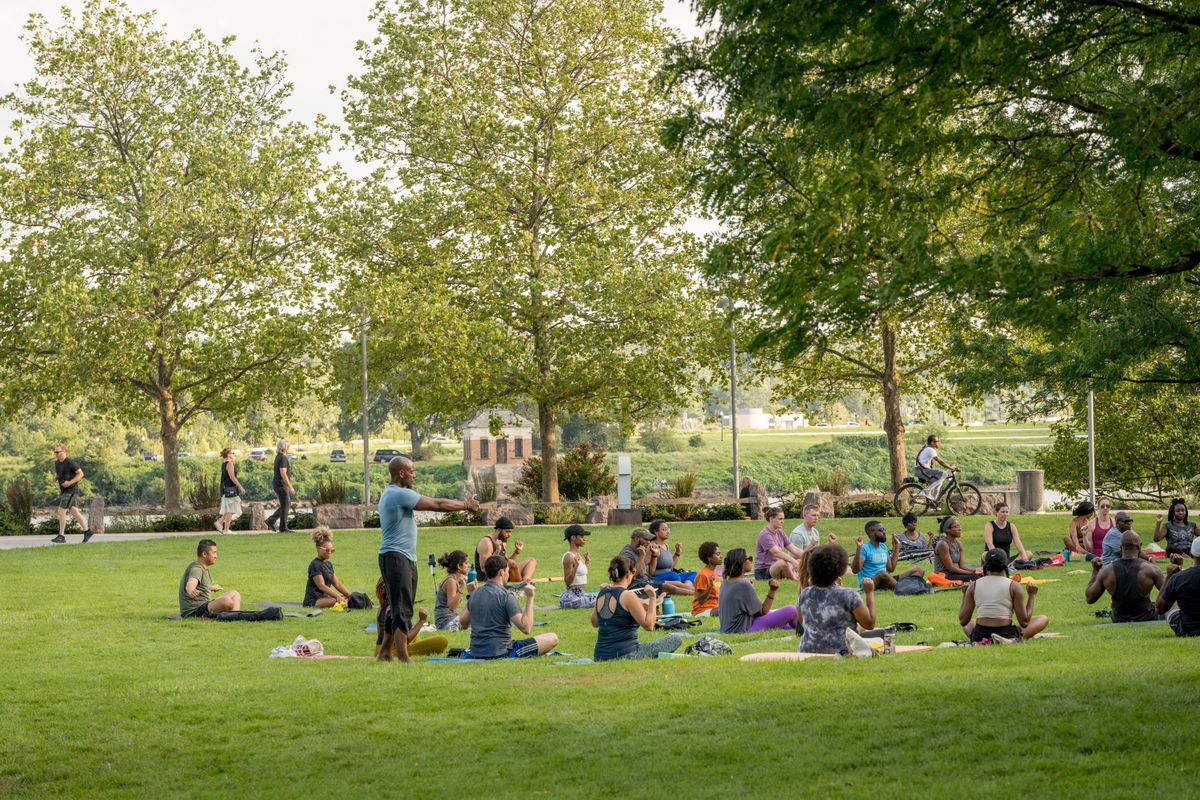 An instructor leading a large outdoor fitness class on a riverside park lawn